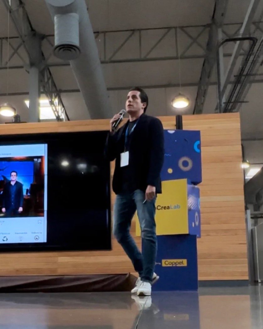 Edgar Temores hablando con micrófono en un escenario, con una pantalla que lo proyecta a su izquierda y un panel azul de patrocinadores detrás