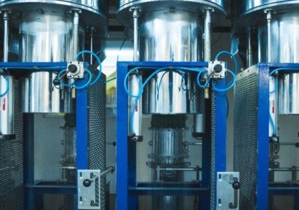 Tres columnas cilíndricas de acero inoxidable con bastidor azul y mangueras neumáticas, en la línea de prensado