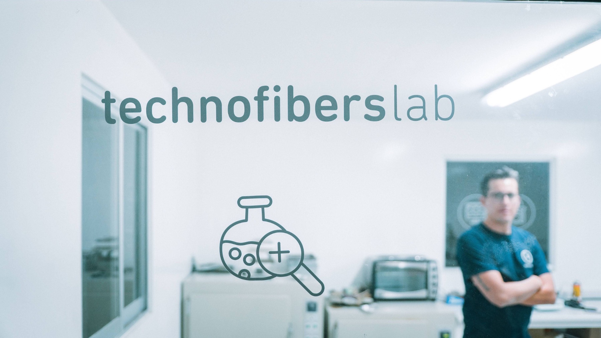 Puerta de cristal del laboratorio de Technofibers, con el rótulo technofibers lab y, detrás y desenfocada, la sala con un técnico de brazos cruzados entre los equipos