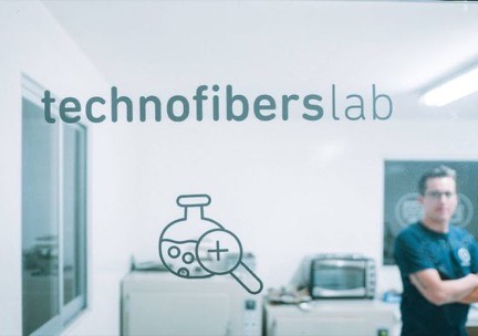 Puerta de cristal rotulada technofibers lab, con un técnico desenfocado al fondo entre equipos de laboratorio