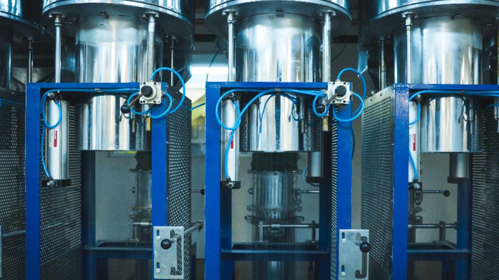 Tres columnas cilíndricas de acero inoxidable sobre bastidores azules, con mangueras neumáticas y rejillas de protección, en la línea de producción de Technofibers